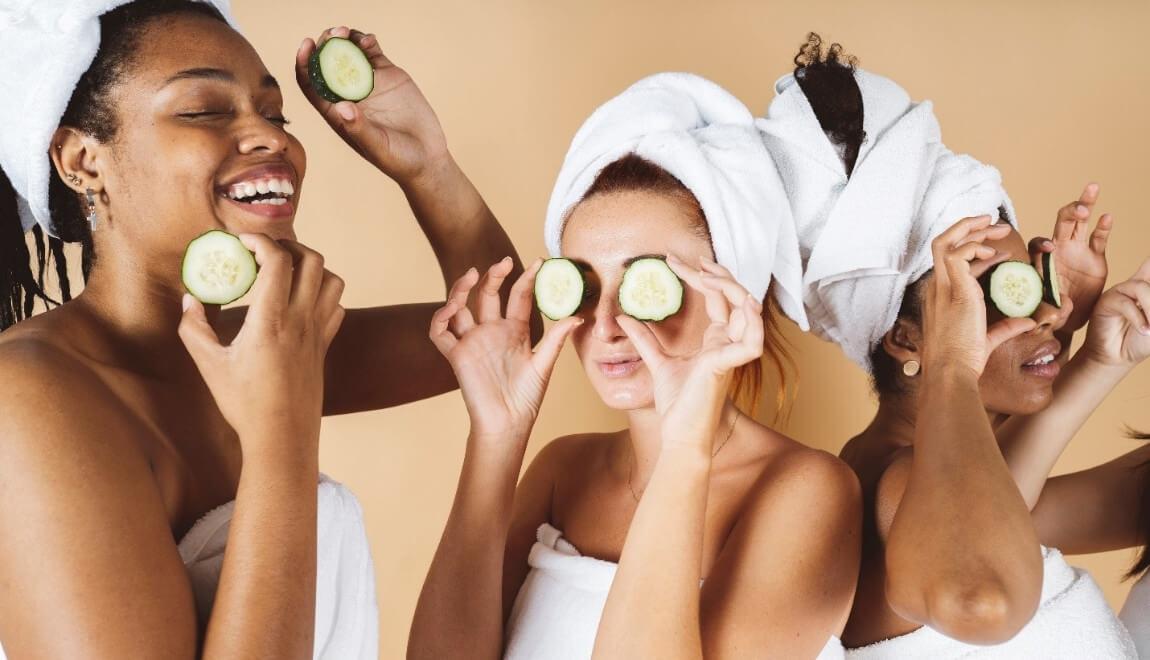 Women wrapped in towels holding slices of cucumbers over their eyes.