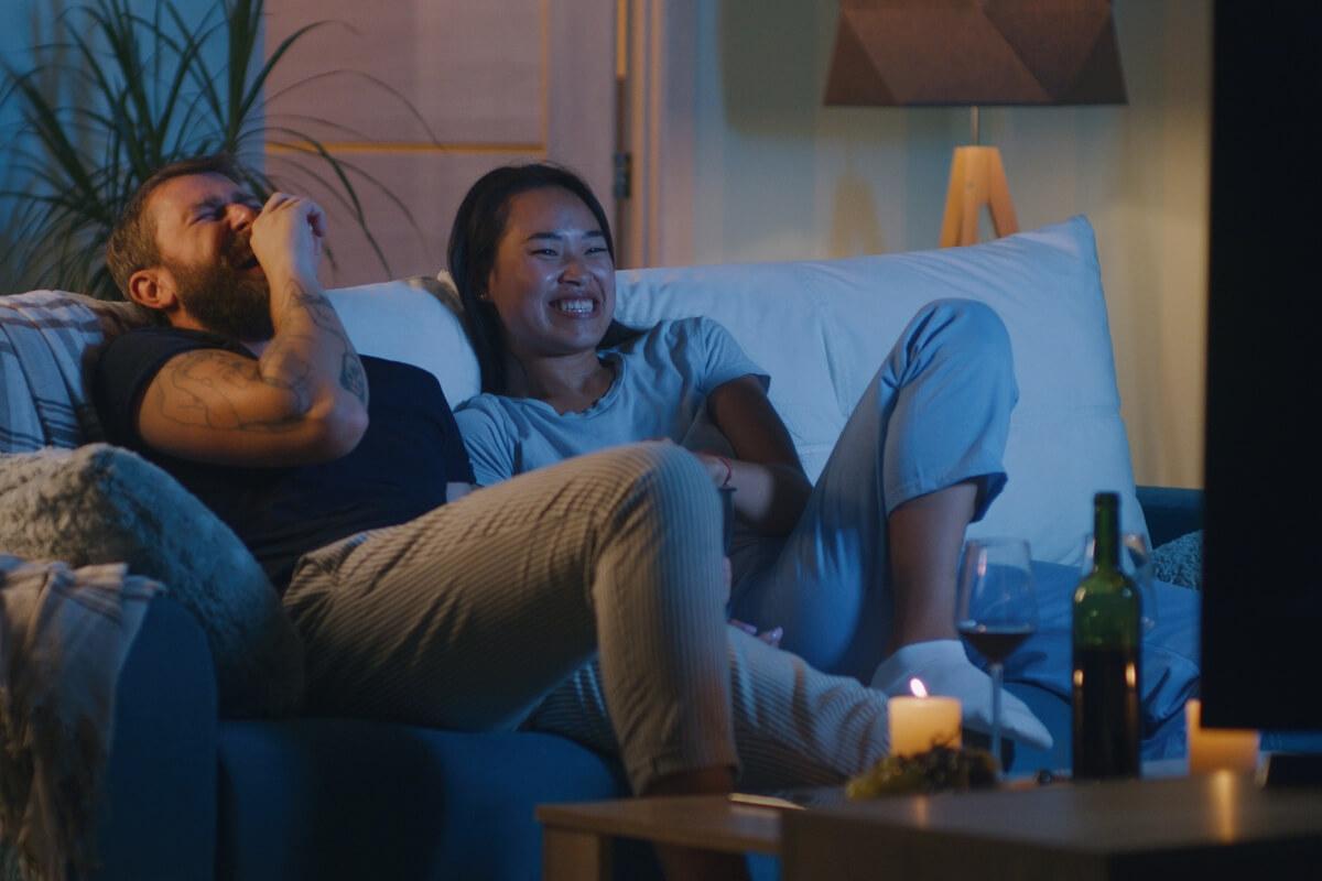 A young couple laughs at a movie on the TV while sitting on a couch.