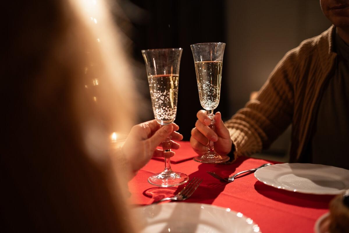 Two people hold glasses of champagne on opposite sides of a romantic dinner table.