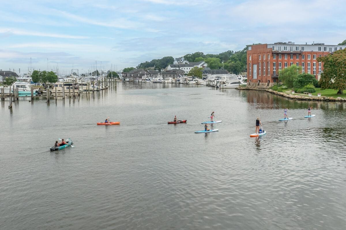 People kayak and paddleboard down the Mystic River with the Mystic Historic District in the background.