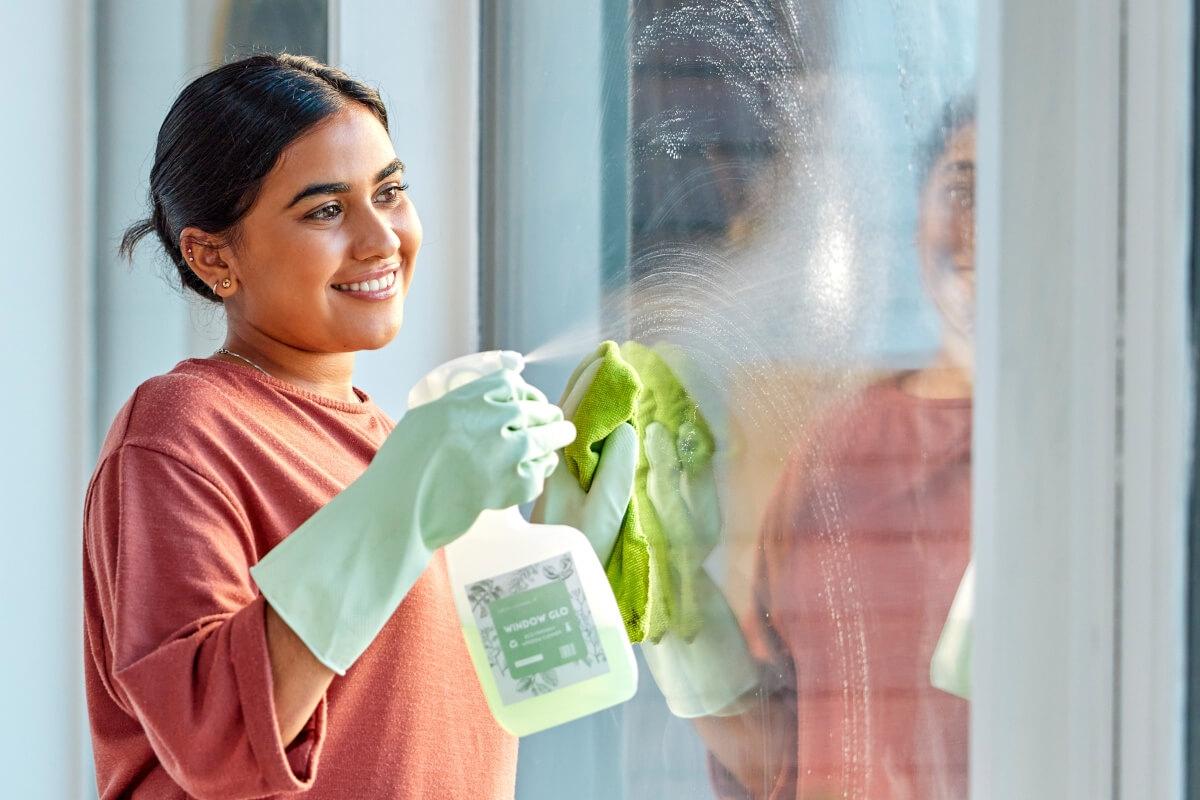 A smiling young woman cleans a window with glass cleaner and a microfiber towel.