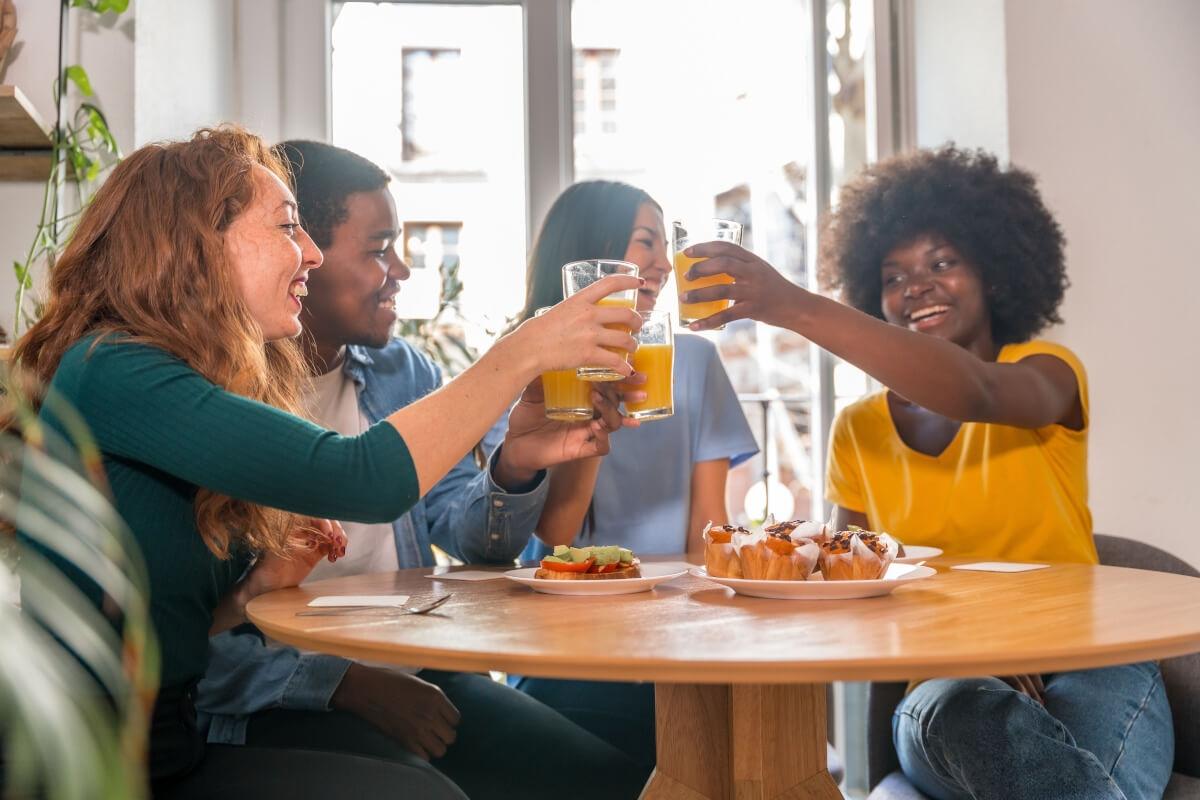 Four friends clink glasses of orange juice while eating breakfast.