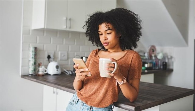 Woman with a cup of coffee in one hand and a smartphone in the other standing in her kitchen.