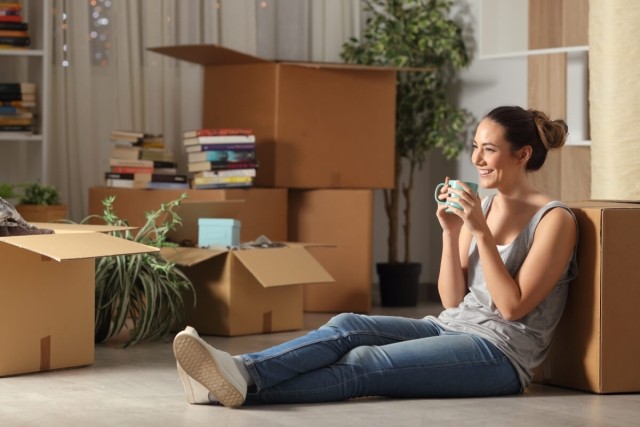 A smiling young woman surrounded by moving boxes sits on the floor of her apartment and sips coffee.