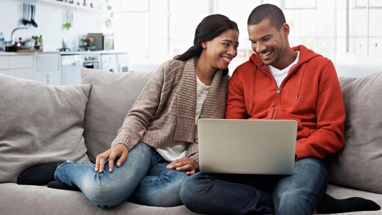 Smiling couple browses apartment listings on laptop while sitting on couch together