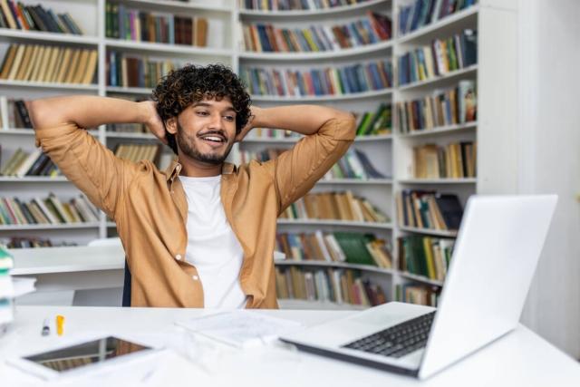 A person sitting back with their hands behind their head watching their laptop with a smile on their face.