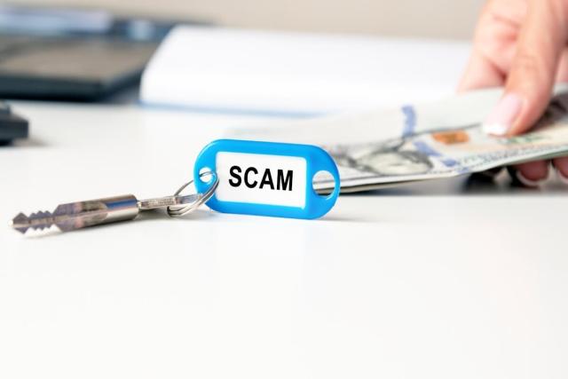 The word "scam" on a blue key fob in front of a hand holding money.