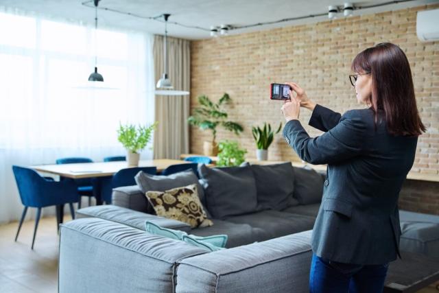 Woman in professional clothing taking photo of rental living room with smartphone.