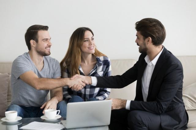 A landlord wearing a suit shakes hands with a young couple looking to rent.