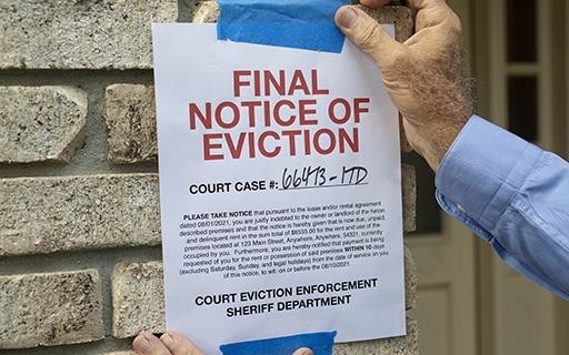 Image of a man taping a document that reads "FINAL NOTICE OF EVICTION" printed in red ink with all capital letters to a brink wall beside a front door entrance