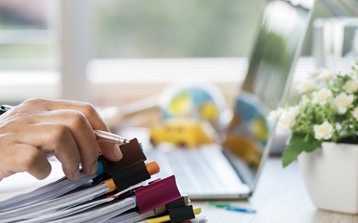 Hand with holding pencil reaches for a stack of documents with binder clips while sitting at a desk with a laptop and plant